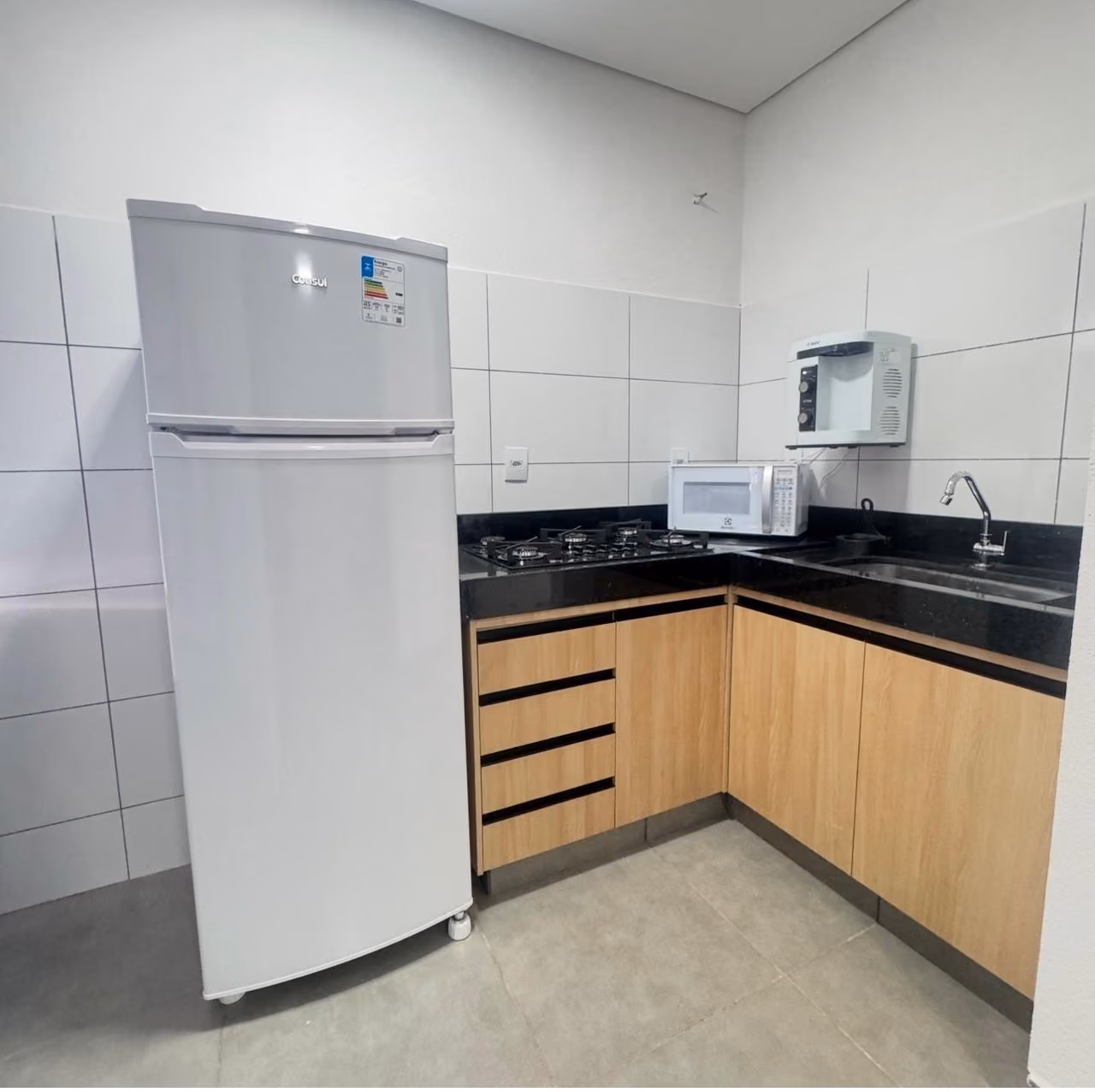 Cozinha compacta com bancada preta, pia de inox, fogão de mesa, armários cinza, geladeira branca, micro-ondas no nicho de prateleira de madeira e decoração com planta verde.