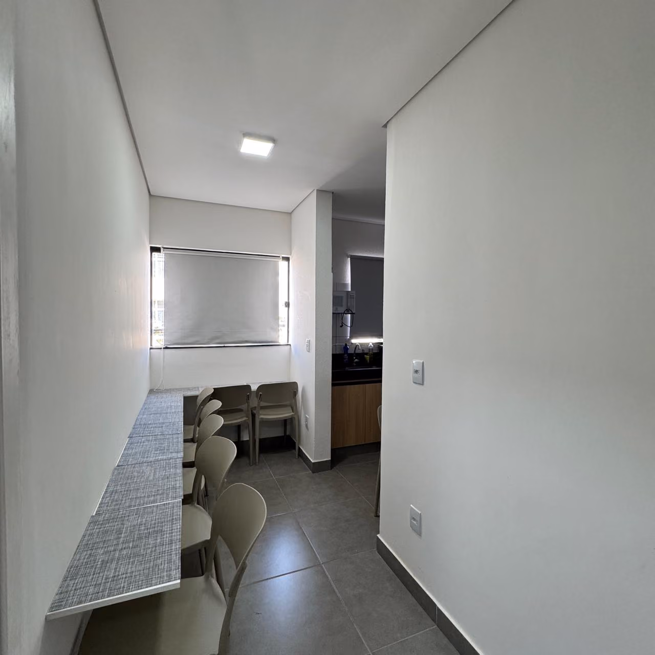 Quarto moderno com cama branca, almofadas bege, cobertor, cadeira de escritório branca e três quadros decorativos abstratos na parede, com janela mostrando prédios ao fundo.